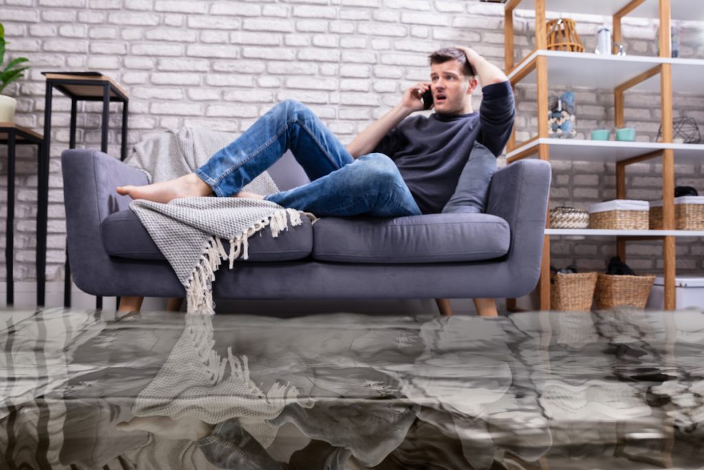 Flooded Floor From Water Leakage In Front Of Man Sitting On Sofa Calling To Plumber